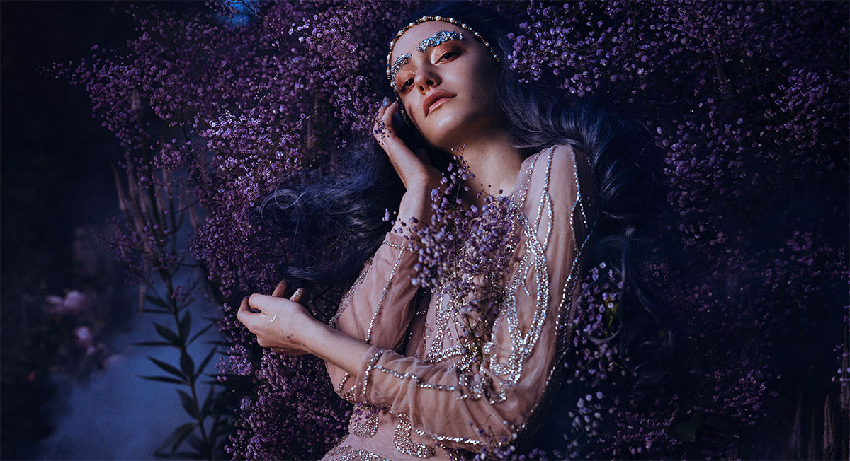 Ethereal woman surrounded by purple flowers wearing beaded clothes and glittery make up taken by Bella Kotak